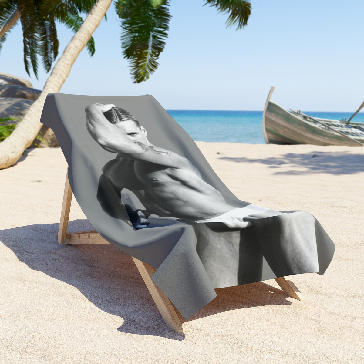 A beach chair on a sandy tropical beach draped with a gray and white towel with a black-and-white photo of a hunky gay man in a bathing suit. Sexy and ready for the beach. For sale in The Pink Rocket New Orleans gay owned gift shop.