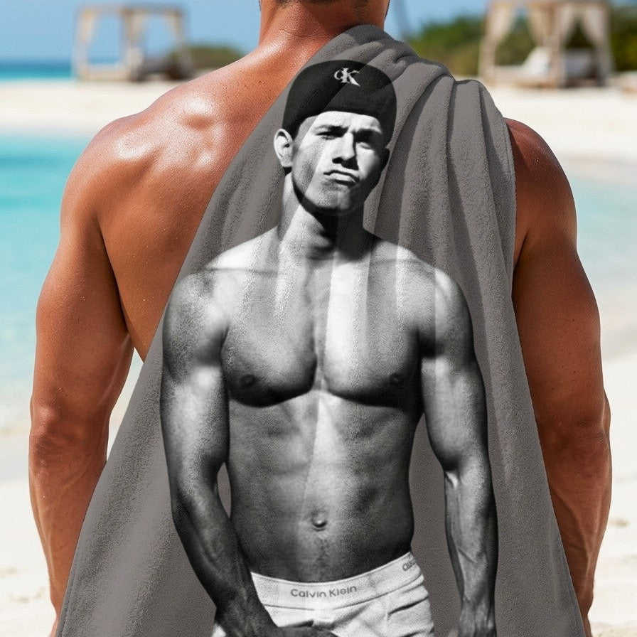 A gray marky mark towel draped over the back of a shirtless gay man at a beach, featuring a black-and-white printed photo of a muscular marky mark wearing a Calvin Klein cap and Calvin Klein underwear. Sold in The Pink Rocket ew Orleans a gay small business.