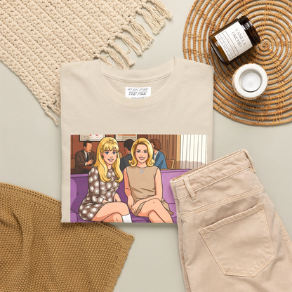 Folded off-white short-sleeve t-shirt with a graphic print of two women, Jeannie and Samantha Stevens sitting on a purple couch with the text 'Cocktail Magic' below, displayed against a solid dark olive green background. Designed and for sale in The Pink Rocket New Orleans gay gift shop.