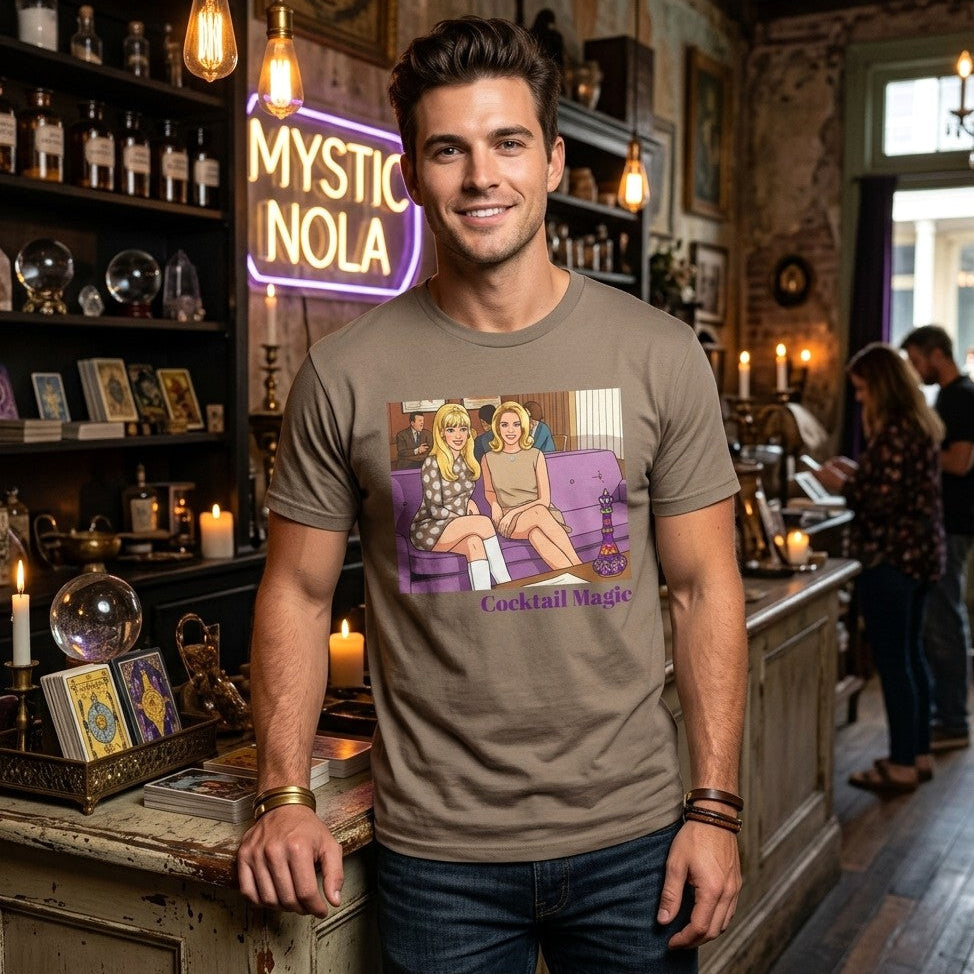 Model wearing a brown short-sleeve t-shirt with a graphic print of two women, Jeannie and Samantha Stevens sitting on a purple couch with the text 'Cocktail Magic' below, displayed against a solid dark olive green background. Designed and for sale in The Pink Rocket New Orleans gay gift shop.
