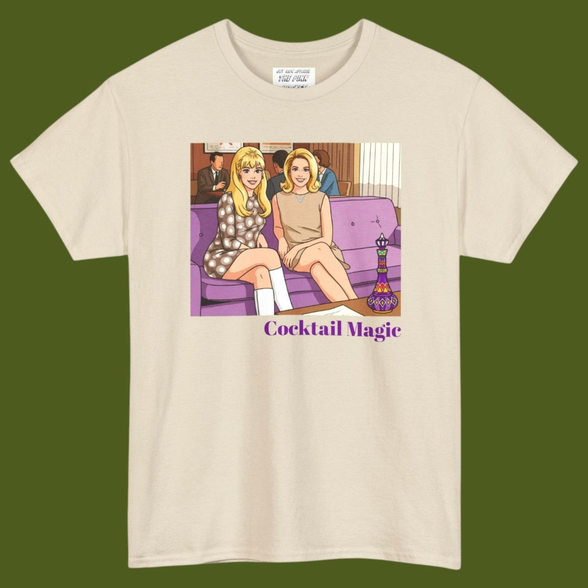 An off-white short-sleeve t-shirt with a graphic print of two women, Jeannie and Samantha Stevens sitting on a purple couch with the text 'Cocktail Magic' below, displayed against a solid dark olive green background. Designed and for sale in The Pink Rocket New Orleans gay gift shop.