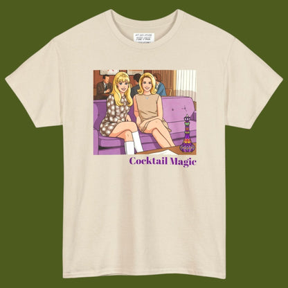 An off-white short-sleeve t-shirt with a graphic print of two women, Jeannie and Samantha Stevens sitting on a purple couch with the text 'Cocktail Magic' below, displayed against a solid dark olive green background. Designed and for sale in The Pink Rocket New Orleans gay gift shop.