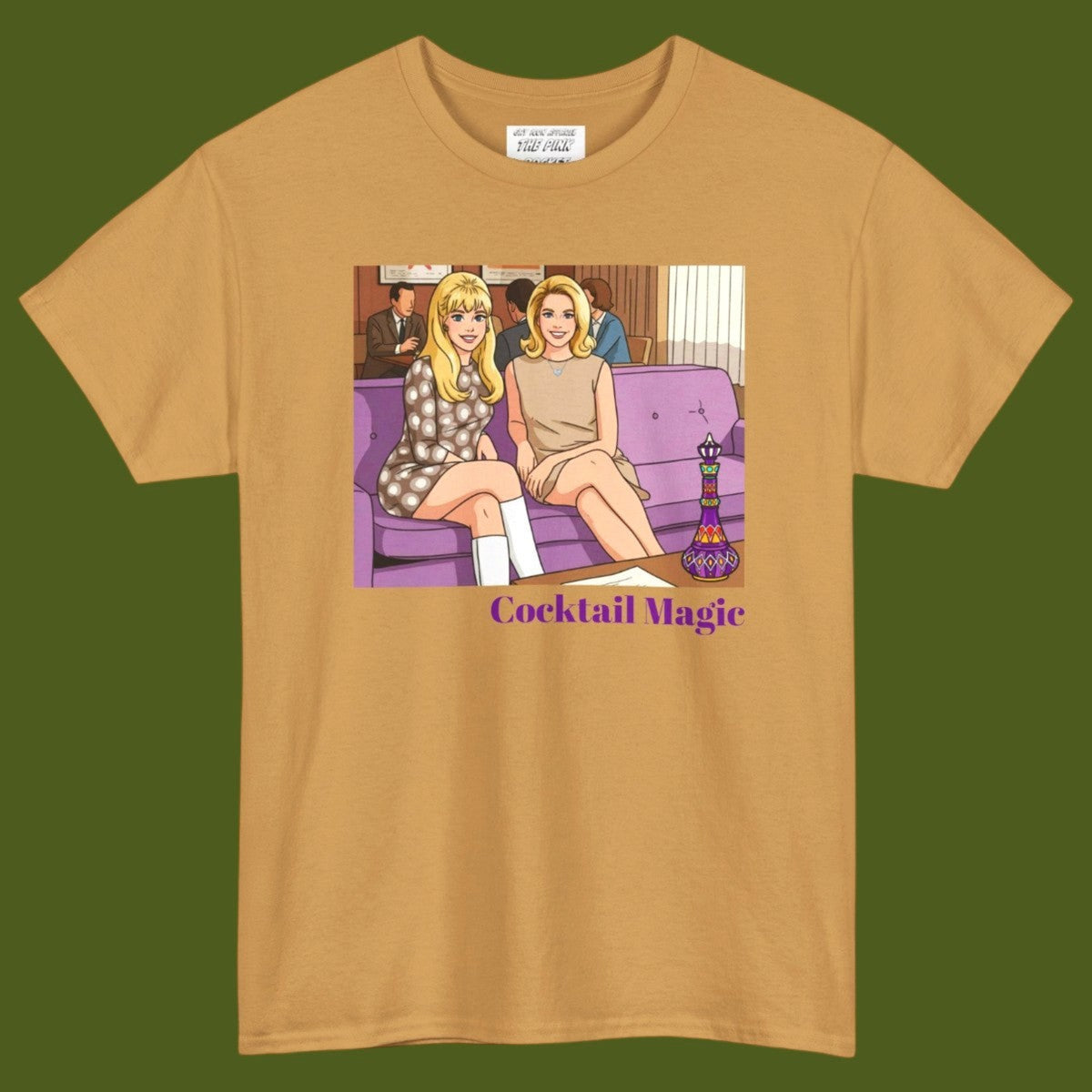An gold short-sleeve t-shirt with a graphic print of two women, Jeannie and Samantha Stevens sitting on a purple couch with the text 'Cocktail Magic' below, displayed against a solid dark olive green background. Designed and for sale in The Pink Rocket New Orleans gay gift shop.