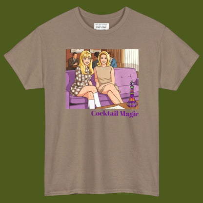 An brown short-sleeve t-shirt with a graphic print of two women, Jeannie and Samantha Stevens sitting on a purple couch with the text 'Cocktail Magic' below, displayed against a solid dark olive green background. Designed and for sale in The Pink Rocket New Orleans gay gift shop.