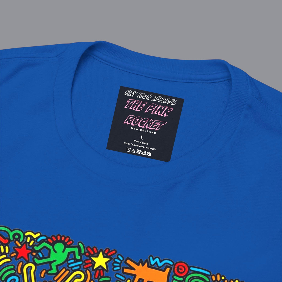 An close up of a blue short-sleeve crewneck t-shirt featuring a colorful graphic of a Keith Haring style 'Gay Icon' graphic, with a red heart and the words 'GAY ICON' in red, displayed against a solid blue background. Sold in The Pink Rocket New Orleans gay gift shop.