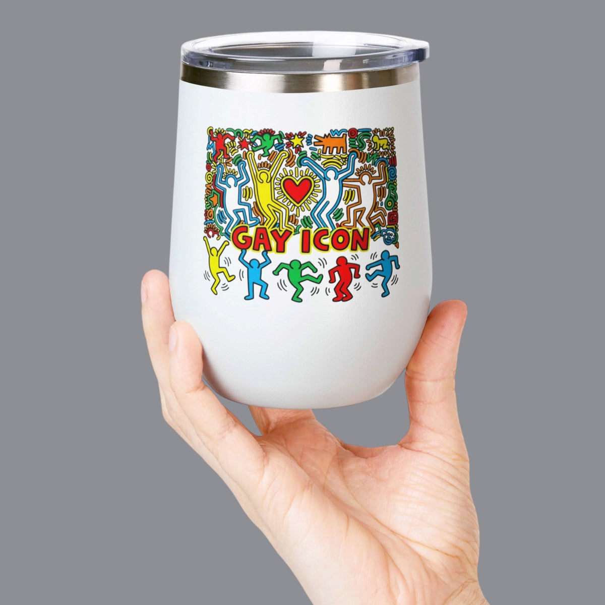 A hand holding a white 12oz. insulated stemless wine tumbler with a clear lid. The tumbler features a colorful graphic of dancing figures and the words 'GAY ICON' in red, yellow, and blue. Designed and sold in The Pink Rocket New Orleans gay gift shop.