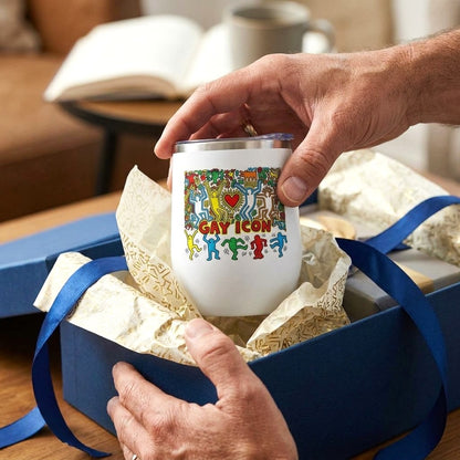 A white 12oz. insulated tumbler with a colorful 'GAY ICON' graphic is being held by a person's hands over an open blue gift box. Designed and sold in The Pink Rocket New Orleans gift shop.