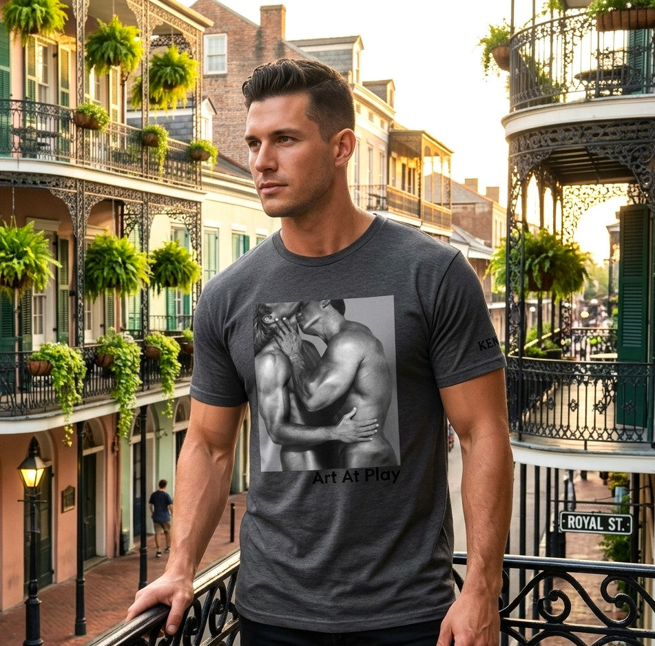 A handsome gay male modeling a heather gray crewneck t-shirt featuring a black and white graphic print of a nude gay male couple embracing and kissing, with the text 'Art At Play' printed below the image. Designed and for sale in The Pink Rocket ew Olreans gay gift shop.