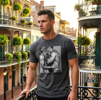 A handsome gay male modeling a heather gray crewneck t-shirt featuring a black and white graphic print of a nude gay male couple embracing and kissing, with the text 'Art At Play' printed below the image. Designed and for sale in The Pink Rocket ew Olreans gay gift shop.