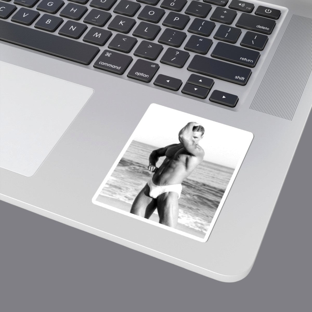 A black and white photograph of a sexy hunky muscular gay man posing on a beach, wearing white briefs. The image is presented with a gray border. For sale in The Pink Rocket New Orleans gay gift shop.