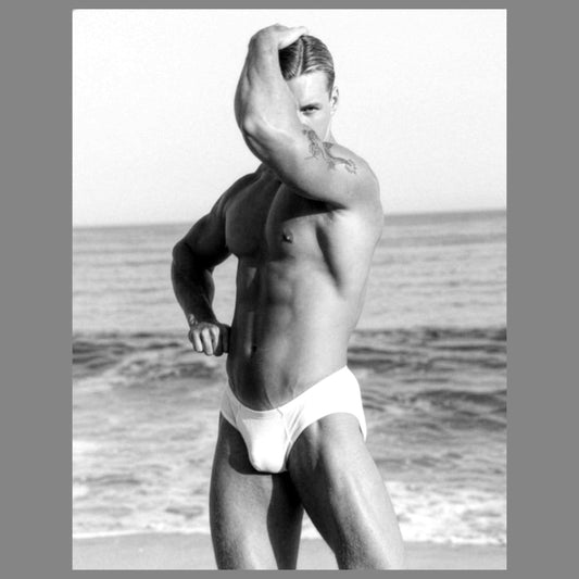 A black and white photograph of a sexy hunky muscular man posing on a beach, wearing white briefs. The image is presented with a gray border. For sale in The Pink Rocket New Orleans gay gift shop.